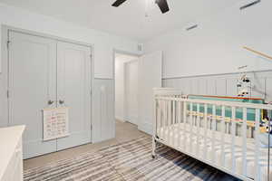 Bedroom with light colored carpet, a wainscoted wall, a crib, a ceiling fan, and a closet