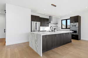 Kitchen featuring dark brown cabinets, stainless steel appliances, wall chimney exhaust hood, light wood-style floors, and light stone counters