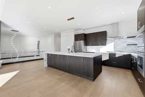 Kitchen featuring modern cabinets, an island with sink, light stone counters, wall chimney range hood, and light wood-style flooring