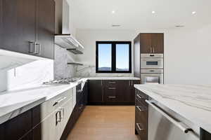 Kitchen with dark brown cabinetry, light stone countertops, modern cabinets, wall chimney exhaust hood, and stainless steel appliances