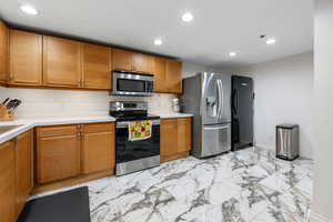 Kitchen with appliances with stainless steel finishes, light countertops, recessed lighting, light marble finish flooring, and tan cabinetry