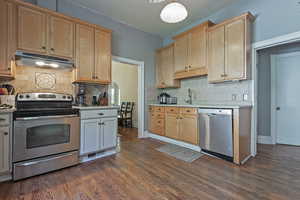 Kitchen featuring stainless steel appliances, light countertops, under cabinet range hood, dark wood-type flooring, and decorative backsplash