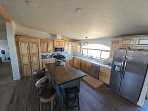 Kitchen featuring stainless steel appliances, vaulted ceiling, light brown cabinets, dark wood-style floors, and a textured ceiling
