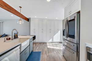 Kitchen featuring stainless steel appliances, light stone countertops, beamed ceiling, white cabinetry, and hanging light fixtures