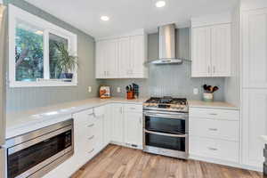 Kitchen with appliances with stainless steel finishes, wall chimney exhaust hood, white cabinets, tasteful backsplash, and light wood-type flooring