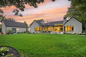 Back of property at dusk featuring a lawn, a chimney, a patio, stone siding, and a shingled roof
