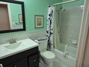 Full bathroom featuring shower / bath combo with shower curtain, vanity, tile walls, light tile patterned floors, and wainscoting
