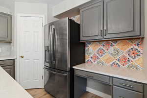 Kitchen featuring gray cabinets, decorative backsplash, light stone counters, stainless steel refrigerator with ice dispenser, and light wood-type flooring