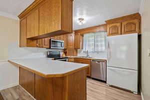 Kitchen with stainless steel appliances, a textured ceiling, light countertops, brown cabinetry, and ornamental molding