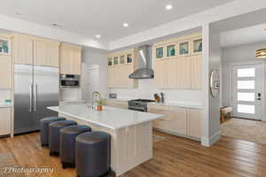 Kitchen with glass insert cabinets, high quality appliances, wall chimney range hood, light brown cabinetry, and light wood-style floors