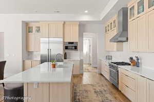 Kitchen featuring glass insert cabinets, light brown cabinetry, wall chimney exhaust hood, light wood-style floors, and recessed lighting