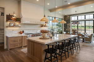 Kitchen featuring open shelves, hanging light fixtures, a breakfast bar area, a center island with sink, and light stone countertops