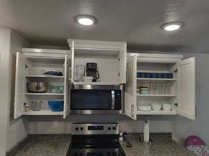 Kitchen view of a textured ceiling, stainless steel appliances, open shelves, white cabinets, and dark stone countertops