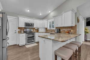 Kitchen with tasteful backsplash, appliances with stainless steel finishes, white cabinets, light stone countertops, and vaulted ceiling