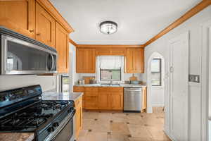 Kitchen featuring stainless steel appliances, arched walkways, crown molding, brown cabinetry, and light tile patterned floors