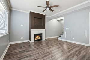 Unfurnished living room with crown molding, dark wood-type flooring, a tiled fireplace, a ceiling fan, and stairs