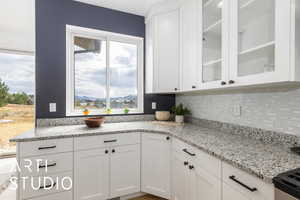 Kitchen with white cabinets, light stone countertops, glass insert cabinets, and tasteful backsplash