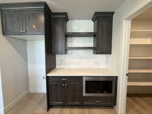 Bar featuring dark brown cabinetry, stainless steel microwave, backsplash, light stone countertops, and light wood-style floors