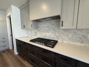 Kitchen with stainless steel gas cooktop, backsplash, premium range hood, dark wood-style flooring, and light stone countertops