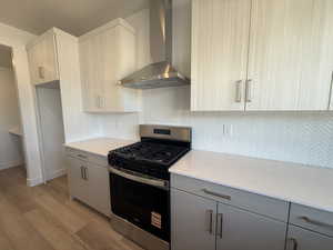 Kitchen with gas range, wall chimney exhaust hood, backsplash, light stone countertops, and light wood-style flooring