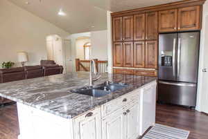 Kitchen featuring stainless steel fridge, arched walkways, dark wood-type flooring, dark stone countertops, and an island with sink