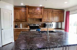 Kitchen with tasteful backsplash, appliances with stainless steel finishes, dark stone counters, brown cabinetry, and recessed lighting
