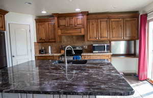 Kitchen featuring dark stone counters, decorative backsplash, appliances with stainless steel finishes, brown cabinetry, and a kitchen island with sink