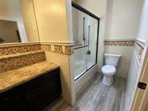 Bathroom featuring tile walls, wainscoting, shower / bath combination with glass door, dark wood-style flooring, and vanity