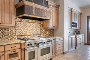 Kitchen with premium range hood, double oven range, decorative backsplash, light stone countertops, and stone tile flooring