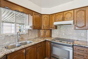 Kitchen featuring stainless steel range with electric stovetop, brown cabinets, light stone countertops, decorative backsplash, and under cabinet range hood