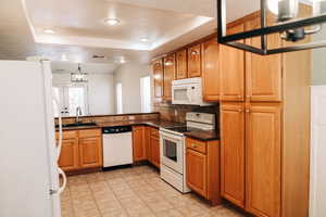 Kitchen with a chandelier, white appliances, a tray ceiling, dark countertops, and brown cabinetry
