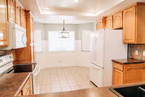 Kitchen featuring white appliances, dark countertops, backsplash, a raised ceiling, and a wainscoted wall