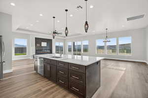Kitchen featuring a raised ceiling, open floor plan, dark brown cabinetry, light wood-style flooring, and decorative light fixtures