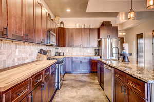 Kitchen featuring pendant lighting, stainless steel appliances, tasteful backsplash, a kitchen island with sink, and light stone counters