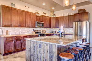 Kitchen featuring decorative backsplash, a kitchen island with sink, stainless steel appliances, light stone countertops, and hanging light fixtures