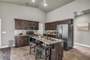 Kitchen with dark brown cabinets, backsplash, appliances with stainless steel finishes, light stone counters, and vaulted ceiling