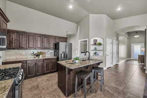 Kitchen featuring dark brown cabinetry, arched walkways, stainless steel appliances, a kitchen bar, and high vaulted ceiling