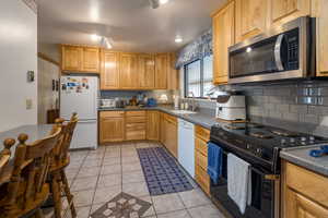 Kitchen featuring white appliances, dark countertops, light tile patterned floors, and backsplash