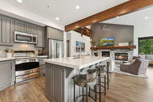 Kitchen with beam ceiling, gray cabinetry, stainless steel appliances, open floor plan, and a stone fireplace