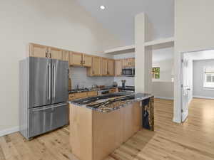 Kitchen featuring dark stone countertops, stainless steel appliances, high vaulted ceiling, light brown cabinetry, and light wood-style floors