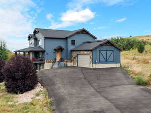 View of front facade with a garage, asphalt driveway, a shingled roof, covered porch, and stone siding