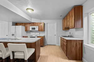 Kitchen featuring brown cabinetry, a kitchen breakfast bar, light countertops, white appliances, and light wood-style floors