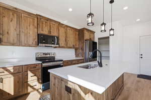 Kitchen with stainless steel appliances, brown cabinetry, an island with sink, light wood finished floors, and decorative light fixtures