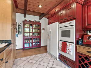 Kitchen with white appliances, dark stone counters, light tile patterned floors, a wooden ceiling with exposed beams, and reddish brown cabinets