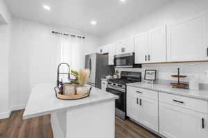 Kitchen with an island with sink, stainless steel appliances, white cabinetry, light stone countertops, and recessed lighting
