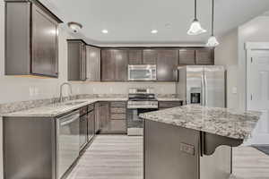 Kitchen with appliances with stainless steel finishes, a kitchen island, decorative light fixtures, light stone counters, and dark brown cabinetry
