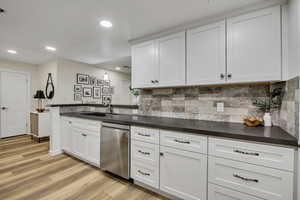 Kitchen with white cabinetry, stainless steel dishwasher, dark countertops, decorative backsplash, and light wood-type flooring