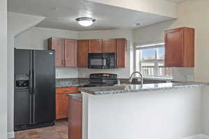 Kitchen featuring black appliances, a peninsula, a textured ceiling, dark stone countertops, and brown cabinetry