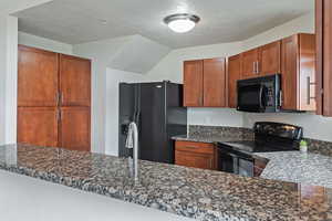 Kitchen with black appliances, a textured ceiling, brown cabinetry, and dark stone counters