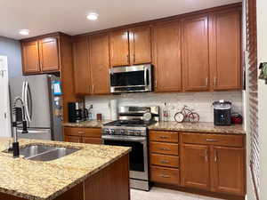 Kitchen featuring appliances with stainless steel finishes, light stone countertops, brown cabinets, decorative backsplash, and recessed lighting